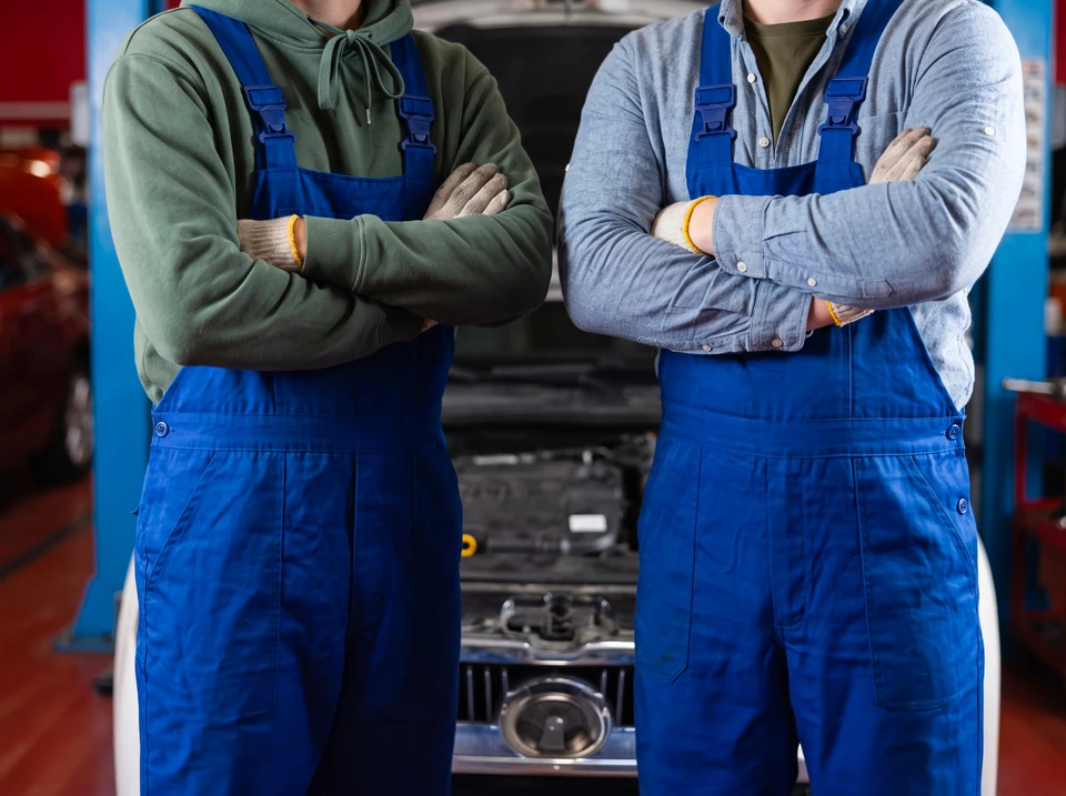 Photographie réaliste en atelier professionnel montrant deux mécaniciens d'âges différents (père et fils) travaillant ensemble sur une moto, penchés sur le moteur ou effectuant une vérification technique. Le père en arrière-plan guidant, le fils au premier plan effectuant l'intervention. Environnement professionnel avec outils, établi, véhicules en arrière-plan. Atmosphère authentique d'apprentissage et de transmission de savoir-faire, éclairage naturel d'atelier industriel.